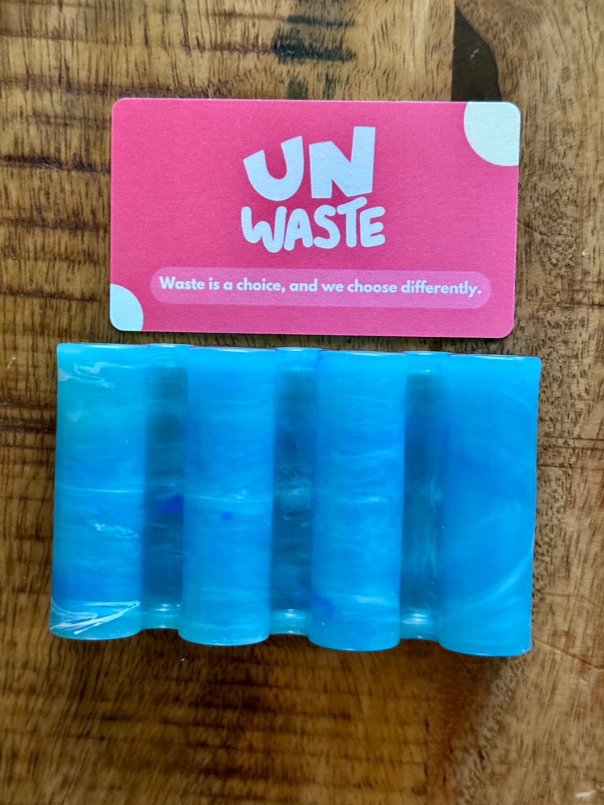 An Upcycled Soap Tray by Unwaste, crafted from blue, translucent recycled plastic with cylindrical shapes, rests on a wooden surface beneath a pink Unwaste card—an ideal addition to any zero-waste bathroom.