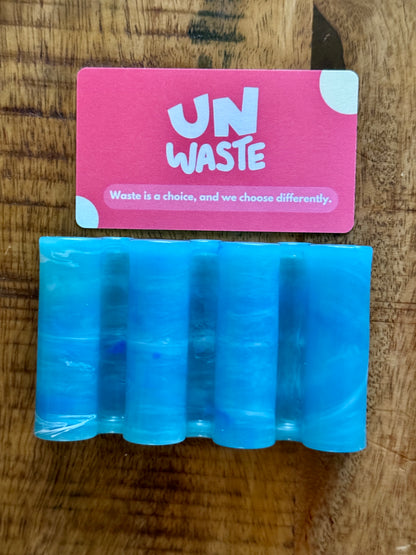 An Upcycled Soap Tray by Unwaste, crafted from blue, translucent recycled plastic with cylindrical shapes, rests on a wooden surface beneath a pink Unwaste card—an ideal addition to any zero-waste bathroom.