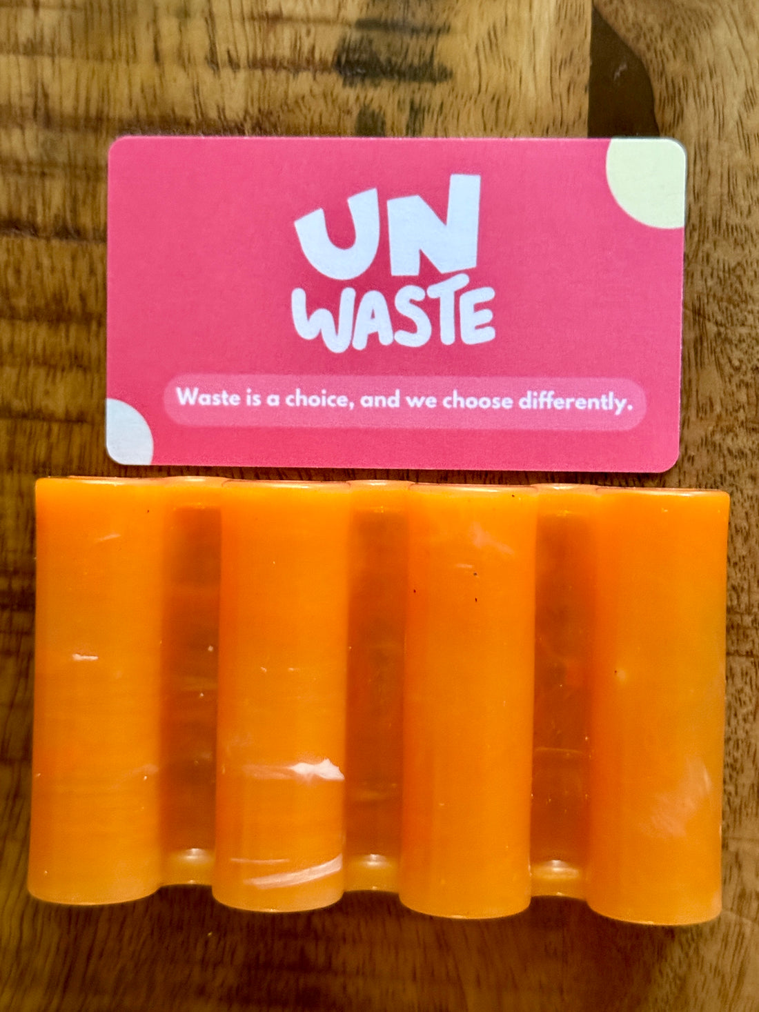 An Upcycled Soap Tray by Unwaste—crafted from recycled plastic in a vibrant orange hand shape—rests on a wooden surface, topped with a pink card that reads “UN WASTE” and the message: “Waste is a choice, and we choose differently.” This inspiring set makes a standout addition to any zero-waste bathroom.