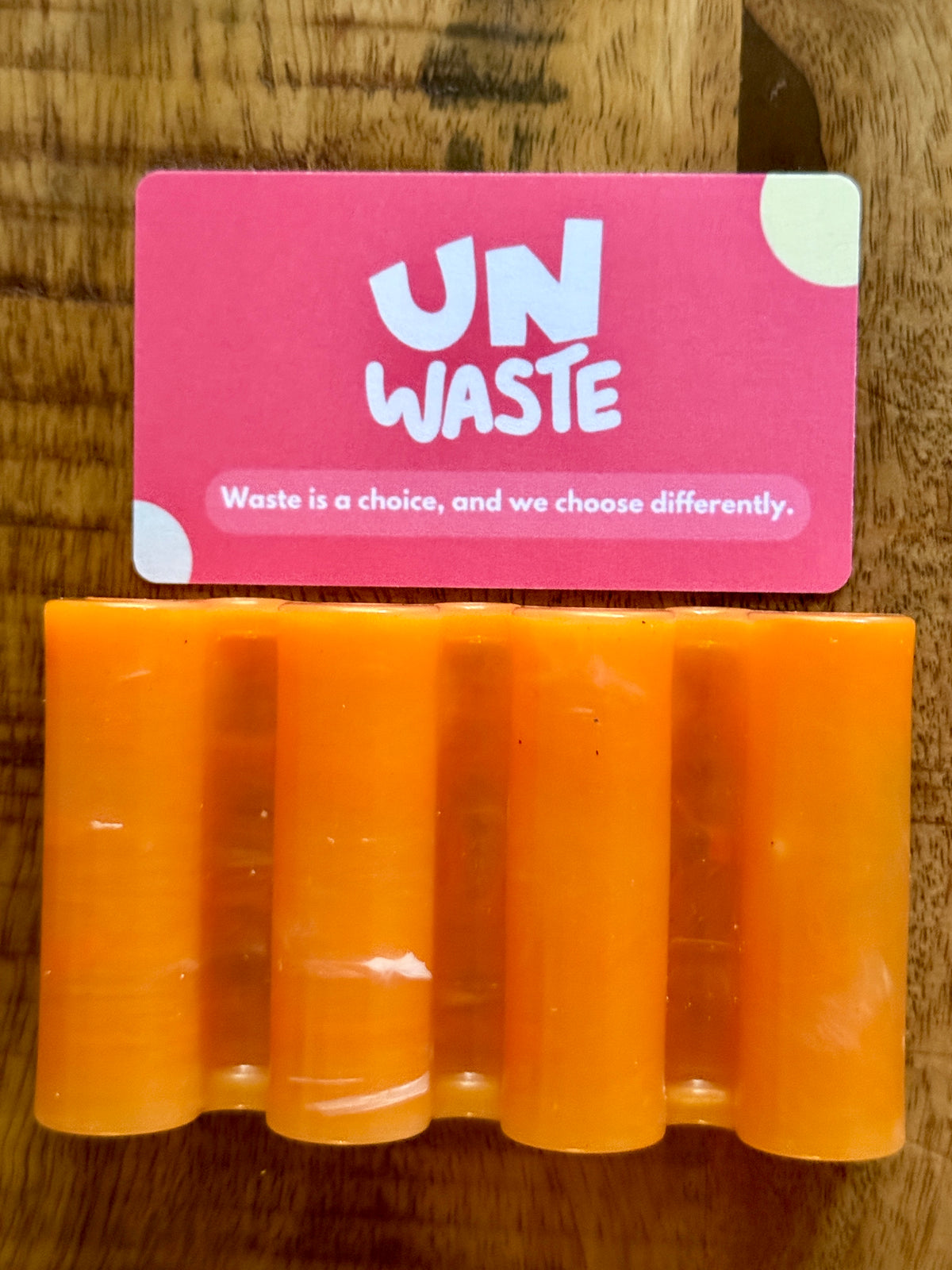 An Upcycled Soap Tray by Unwaste—crafted from recycled plastic in a vibrant orange hand shape—rests on a wooden surface, topped with a pink card that reads “UN WASTE” and the message: “Waste is a choice, and we choose differently.” This inspiring set makes a standout addition to any zero-waste bathroom.