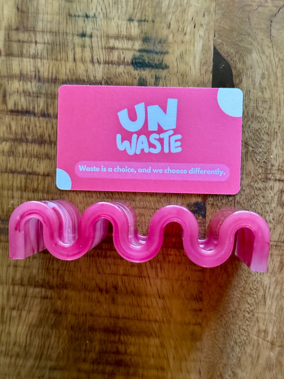 An Upcycled Soap Tray from Unwaste, in a pink wavy hand shape, rests beneath a pink UN WASTE card declaring “Waste is a choice.” Together, they champion a zero-waste bathroom lifestyle on a wooden surface.