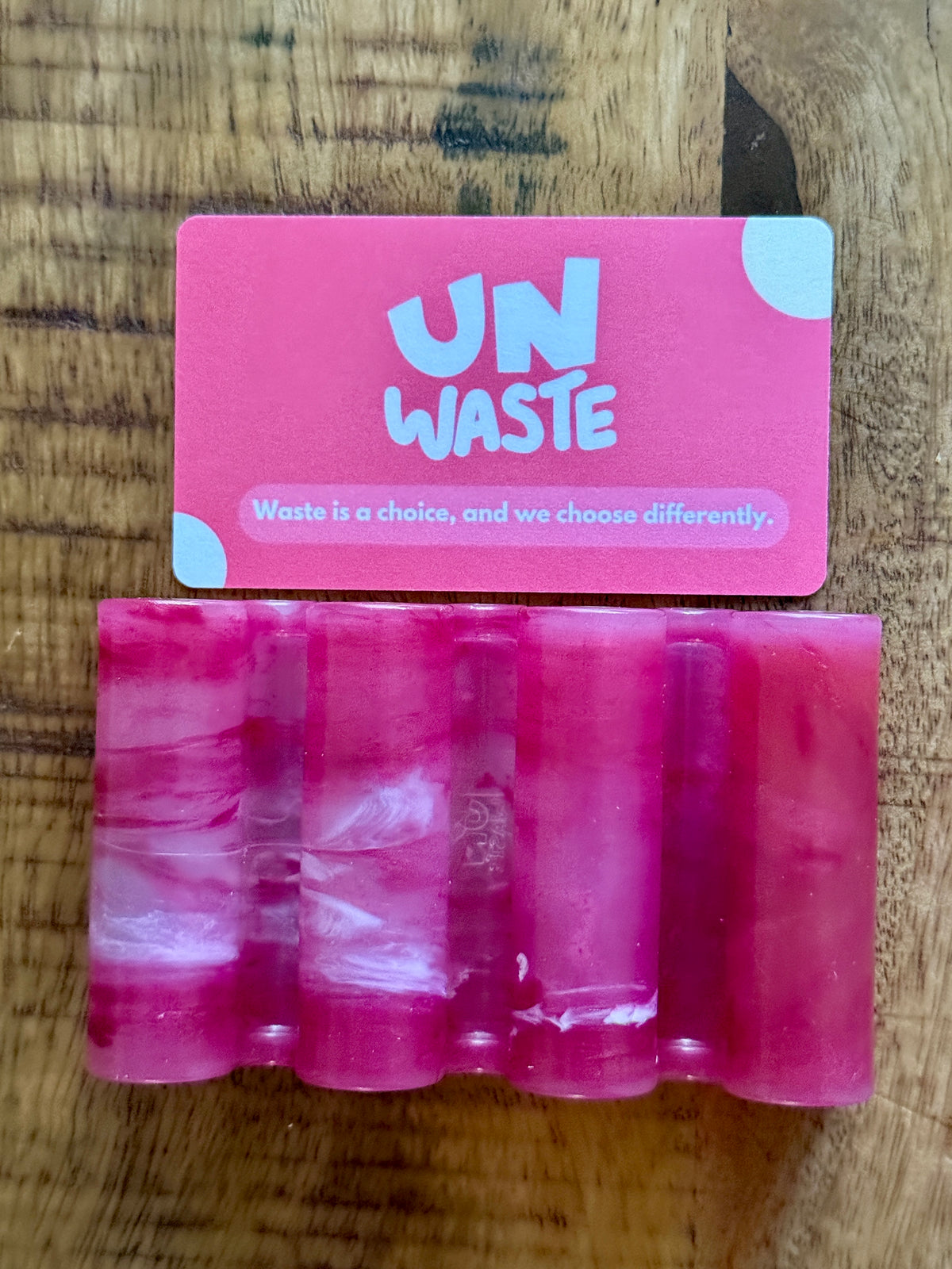 A pink and white marbled soap bar rests on a wooden surface, displayed on the Unwaste Upcycled Soap Tray—a recycled plastic holder—beneath a pink card with “UNWASTE” written on it. Waste is a choice, and we choose differently—bringing beauty to your zero-waste bathroom.