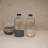 Two 16oz Boston round glass bottles from Waste Less Shop sit on a beige surface with black caps; one has a gray Silicone Boot at its base. Three smooth gray stones are stacked nearby, all set against a neutral background.
