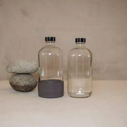 Two 16oz Boston round glass bottles from Waste Less Shop sit on a beige surface with black caps; one has a gray Silicone Boot at its base. Three smooth gray stones are stacked nearby, all set against a neutral background.