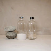 Two stacked smooth gray stones sit beside two clear 16oz Waste Less Shop Boston round bottles with metal caps and silicone boots, all on a neutral surface against a plain beige background.