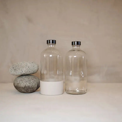 Two stacked smooth gray stones sit beside two clear 16oz Waste Less Shop Boston round bottles with metal caps and silicone boots, all on a neutral surface against a plain beige background.