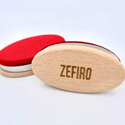 Two Zefiro Fuzz Removers with wooden handles and red fabric surfaces are pictured. One displays the engraved Zefiro brand name on the wooden side, while the other features the red lint-removing surface. Both reusable, eco-friendly Fuzz Removers rest against a plain, light-colored background.