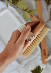 A hand holds the Zefiro Dry Brush, featuring light natural bristles and a fabric strap. Green leaves and another brush appear blurred in the background on a white surface, making it ideal for gentle exfoliation.