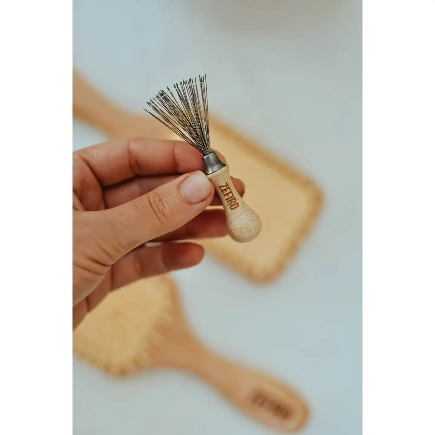 A hand holds the Zefiro Hairbrush Cleaning Tool, a small, plastic-free tool with thin metal bristles. Two eco-friendly wooden hairbrushes sit on a light surface in the blurred background.