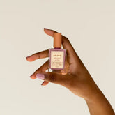 A hand with light purple nails holds a bottle of BKind Nail Polish by BKind against a plain light background.