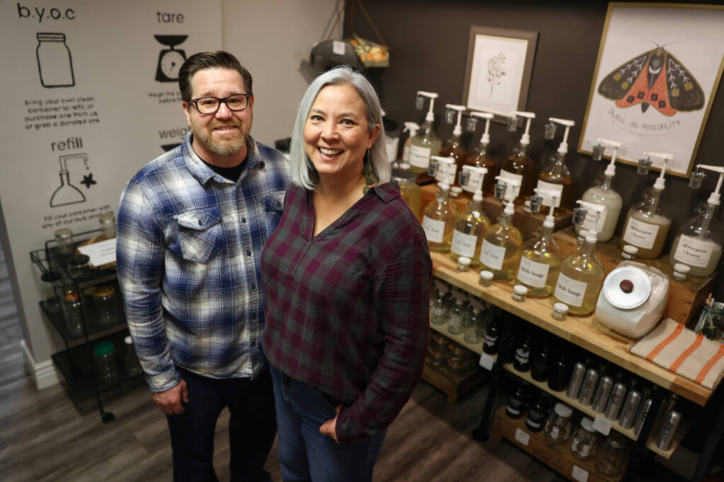 A smiling man and woman stand in a refill store, surrounded by labeled glass dispensers, jars, and eco-friendly products on shelves. The background features framed art and a scale.