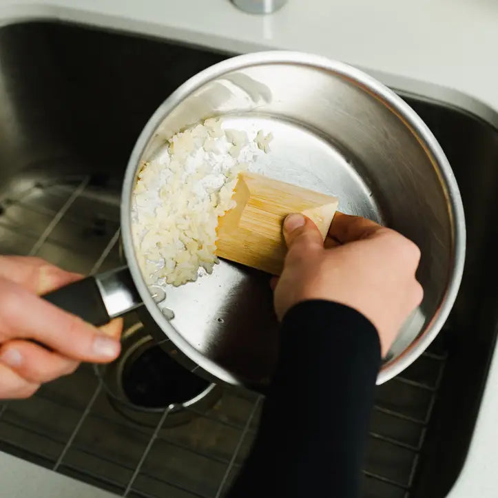 Using the Bamboo Switch Bamboo Pot Scraper, a person scrapes leftover cooked rice from a metal pot into the sink, choosing sustainable, plastic-free cleaning with this eco-friendly kitchen tool.