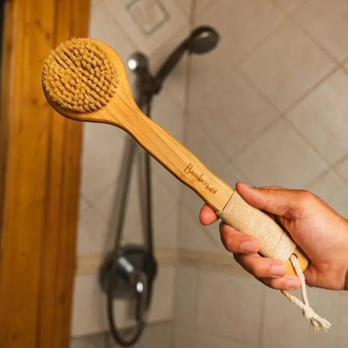 A hand holds the Bamboo Switch Vegan Bamboo Dry Brush with sisal bristles and rope loop in a tiled bathroom with shower and wooden door—your essential exfoliating beauty tool.