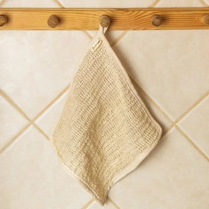 A beige Zefiro Sisal Exfoliating Towel hangs by its loop from a wooden peg on a tiled bathroom wall.