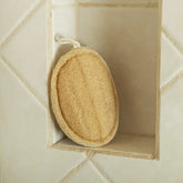 A round, beige Bamboo Switch Loofah with a white hanging loop sits on a recessed shelf in a tiled shower wall with light grout.