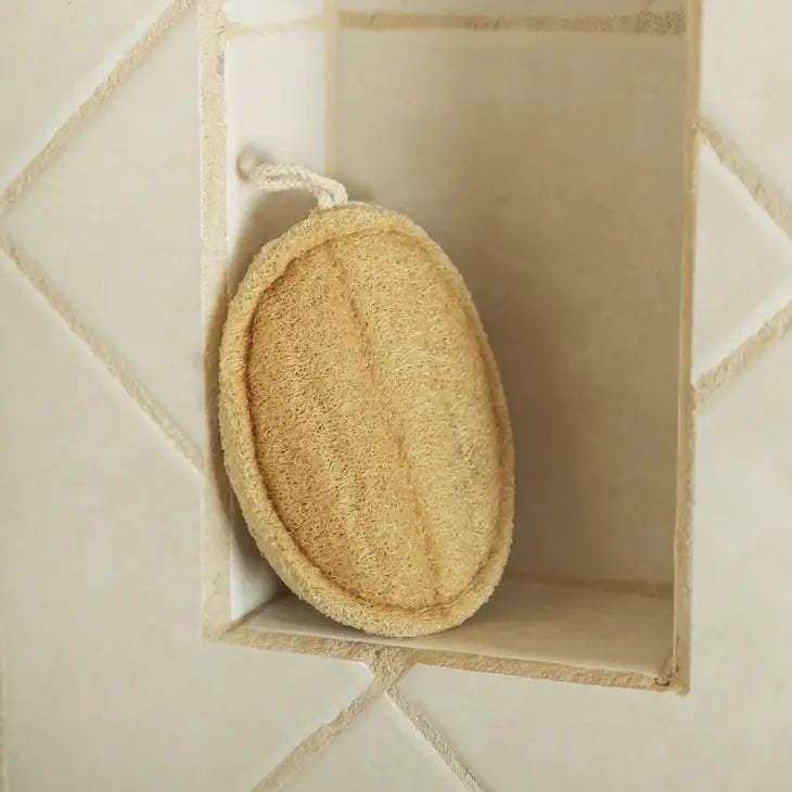 A round, beige Bamboo Switch Loofah with a white hanging loop sits on a recessed shelf in a tiled shower wall with light grout.