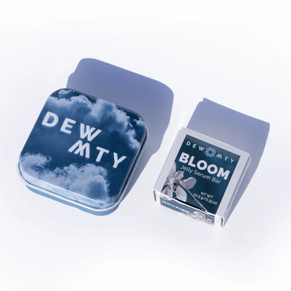 A square tin with a blue sky and clouds design labeled Dewmighty sits next to a box labeled Bloom Skin Serum Bar—an omega-rich serum for skin barrier repair—on a white surface, both casting soft shadows.