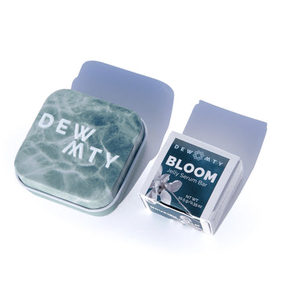 A square greenish-blue Dewmighty tin sits beside a box labeled Bloom Skin Serum Bar—an omega-rich jelly serum for skin barrier repair—on a white background with soft shadows.