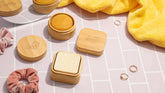 Two Dip Bamboo Travel Cases with Coasters hold round solid soap, arranged with a yellow towel, pink scrunchies, and gold rings on pink tiles. The engraved lid of one case is off, showing the soap inside.