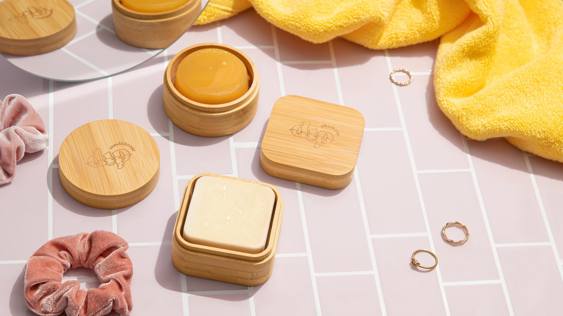 Two Dip Bamboo Travel Cases with Coasters hold round solid soap, arranged with a yellow towel, pink scrunchies, and gold rings on pink tiles. The engraved lid of one case is off, showing the soap inside.