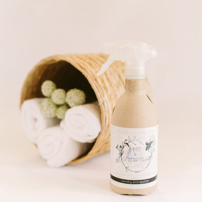 A beige bottle of Laundry Pre-Spotter Stain Remover with a white label, placed in front of a wicker basket containing white towels and laundry balls. The bottle has a foaming trigger spray top.